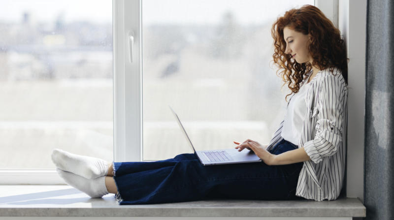 Eine junge Frau sitzt entspannt auf einer Fenserbank mit einem Notebbok auf den Knien.